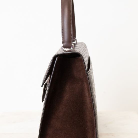 A dark brown handbag shown from a side profile view, featuring a textured crocodile-embossed leather body, a smooth suede-like front panel, and a matching top handle, set against a plain white background.