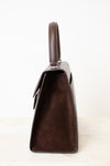 A dark brown handbag shown from a side profile view, featuring a textured crocodile-embossed leather body, a smooth suede-like front panel, and a matching top handle, set against a plain white background.