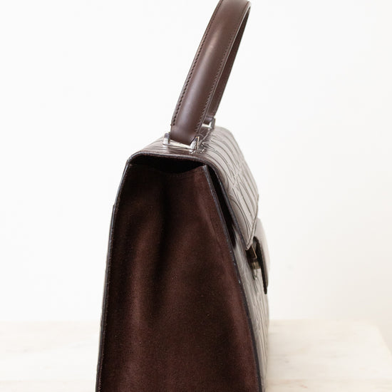 A dark brown handbag shown from a side profile view, featuring a dark brown suede-like interior and a textured dark brown exterior, set against a plain white background.