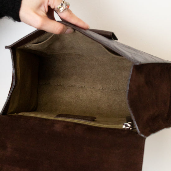 A close-up of a dark brown suede or leather clutch bag being opened by a hand, showing the interior lining and zipper.