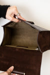 A close-up of a dark brown suede or leather clutch bag being opened by a hand, showing the interior lining and zipper.