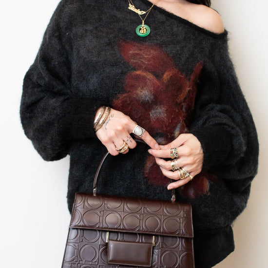 A woman wearing a fuzzy off-the-shoulder black sweater with a large dark red floral print, holding a brown embossed leather handbag. She is wearing gold jewelry, including a green pendant necklace and rings, against a plain off-white background.