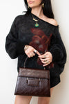 A woman wearing a fuzzy off-the-shoulder black sweater with a large dark red floral print, holding a brown embossed leather handbag. She is wearing gold jewelry, including a green pendant necklace and rings, against a plain off-white background.