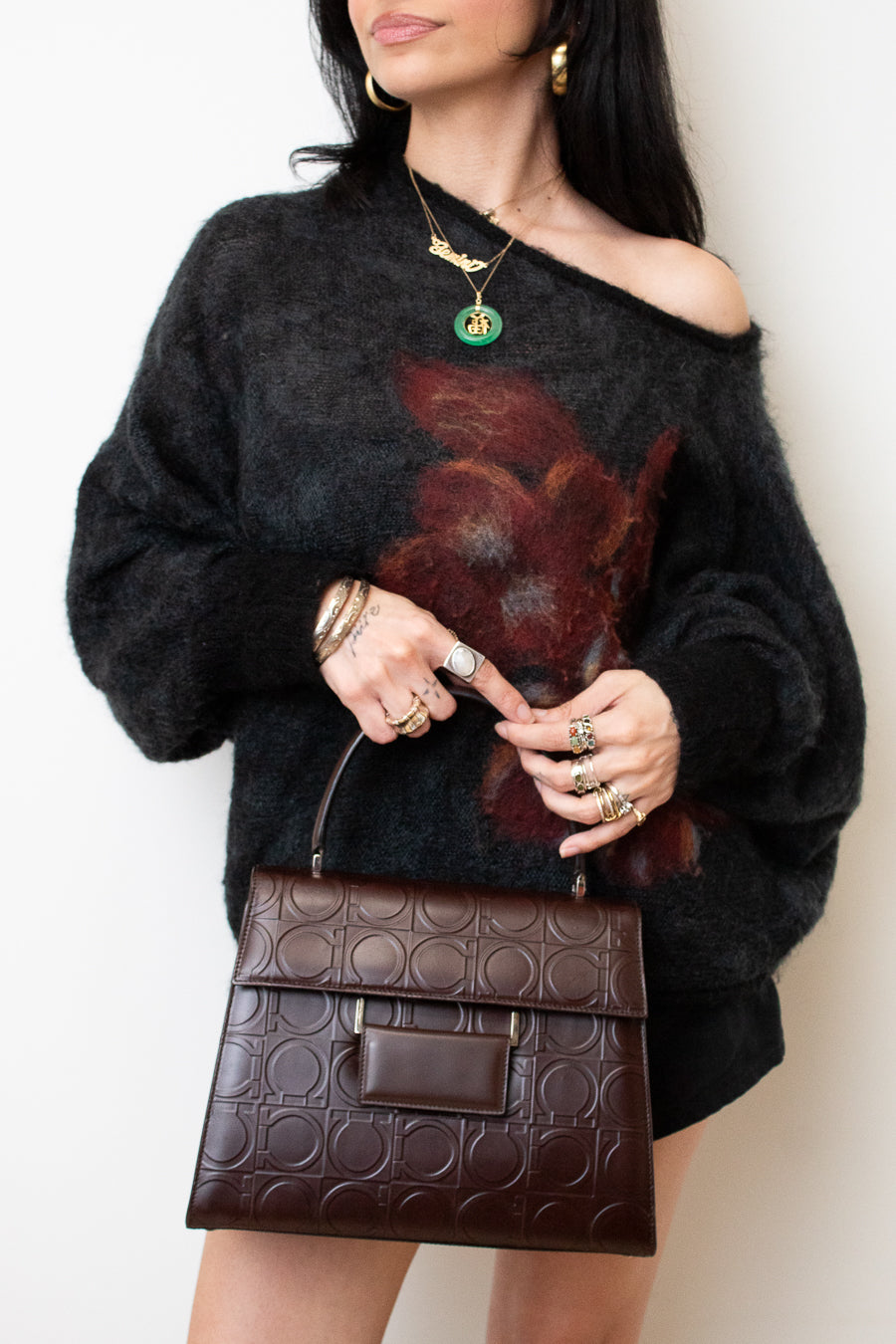 A woman wearing a fuzzy off-the-shoulder black sweater with a large dark red floral print, holding a brown embossed leather handbag. She is wearing gold jewelry, including a green pendant necklace and rings, against a plain off-white background.