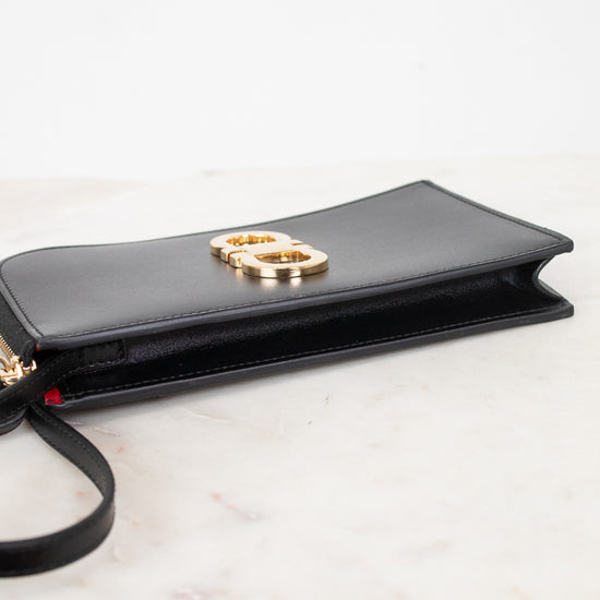 A black leather clutch bag with a gold-tone interlocking logo on the front, resting on a white marble surface against a white background.