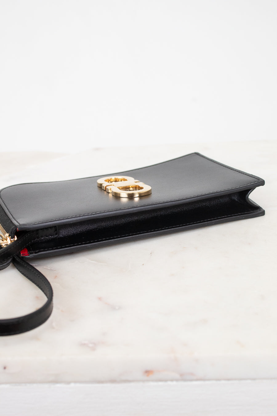 A black leather clutch bag with a gold-tone interlocking logo on the front, resting on a white marble surface against a white background.