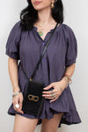 A woman wearing a dark purple puff-sleeve romper and holding a black crossbody bag with a gold-tone FF logo clasp, photographed against a plain light gray background.