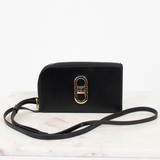 A black leather crossbody wallet with a gold-tone interlocking H-ring clasp, resting on a white marble surface against a white background.