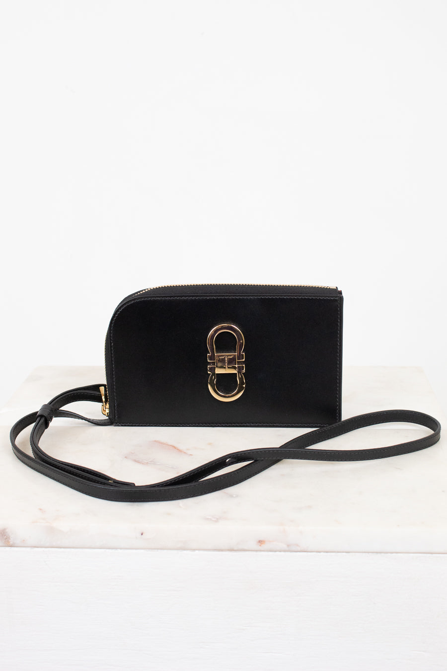 A black leather crossbody wallet with a gold-tone interlocking H-ring clasp, resting on a white marble surface against a white background.