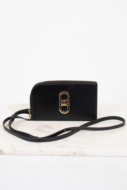 A black leather crossbody wallet with a gold-tone interlocking H-ring clasp, resting on a white marble surface against a white background.