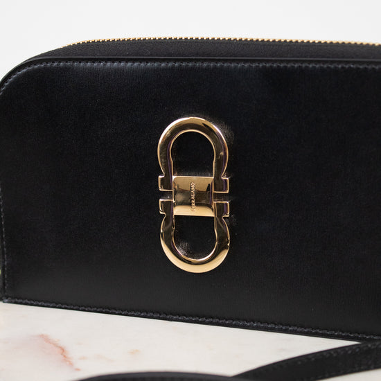A close-up of a black leather shoulder bag featuring a prominent gold-tone metal clasp on the front, resting on a light marble surface with a white background.