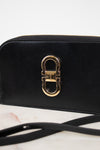 A close-up of a black leather shoulder bag featuring a prominent gold-tone metal clasp on the front, resting on a light marble surface with a white background.