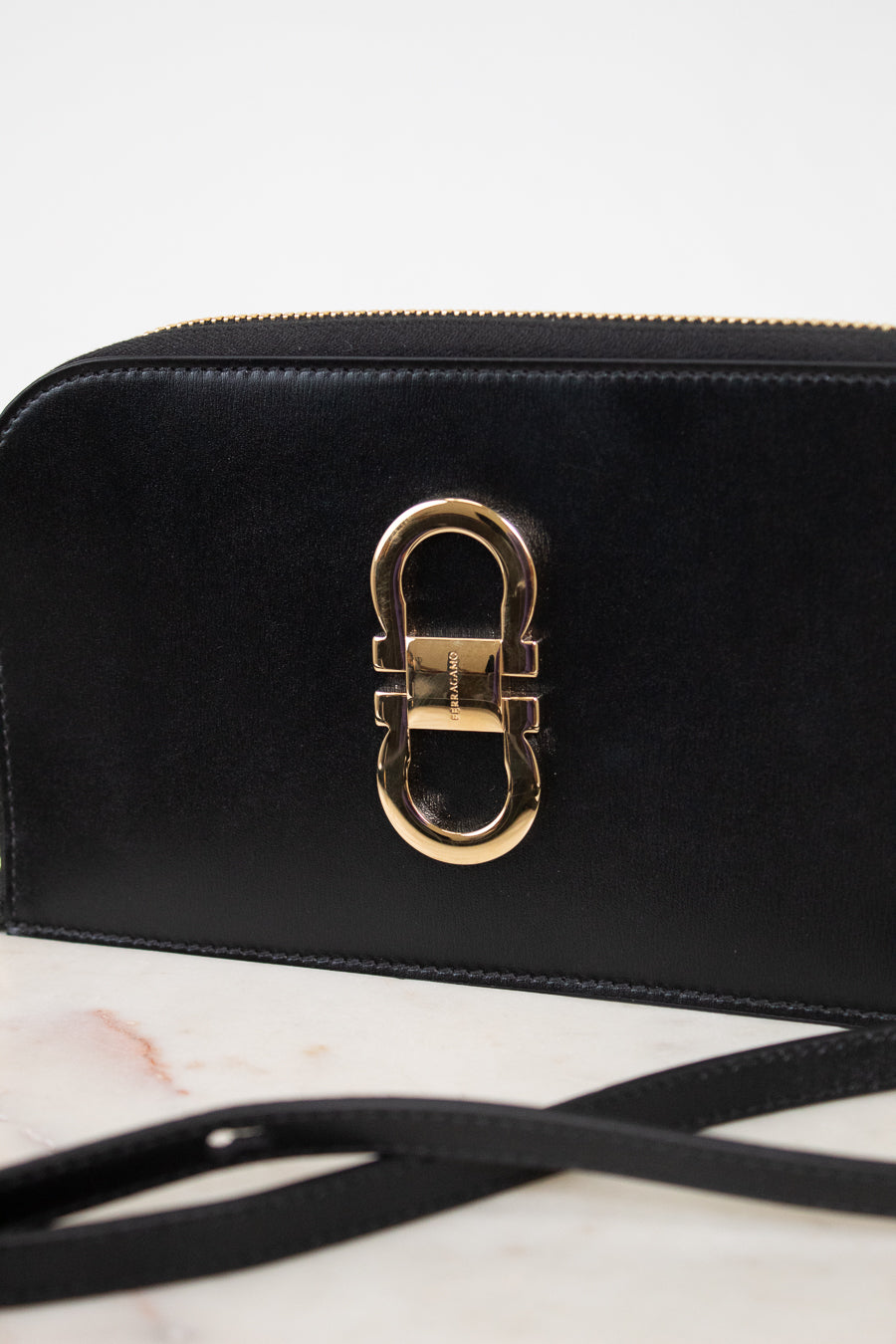 A close-up of a black leather shoulder bag featuring a prominent gold-tone metal clasp on the front, resting on a light marble surface with a white background.