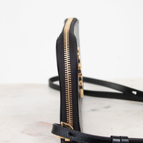 A close-up side view of a black leather crossbody bag with gold-tone hardware and a thin black strap, resting on a light marble surface against a white background.