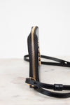 A close-up side view of a black leather crossbody bag with gold-tone hardware and a thin black strap, resting on a light marble surface against a white background.