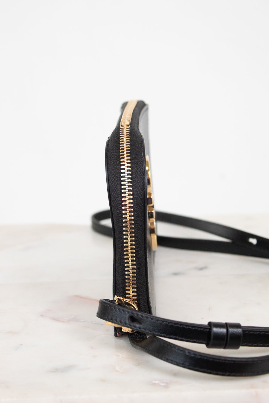 A close-up side view of a black leather crossbody bag with gold-tone hardware and a thin black strap, resting on a light marble surface against a white background.