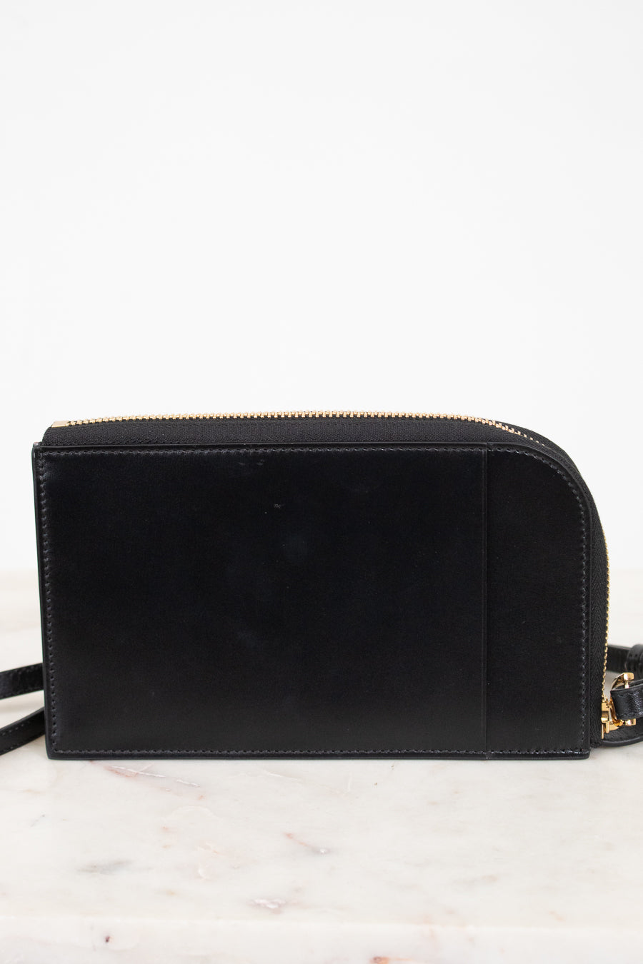 A black leather crossbody bag with gold-tone hardware, resting on a white marble surface against a white background.