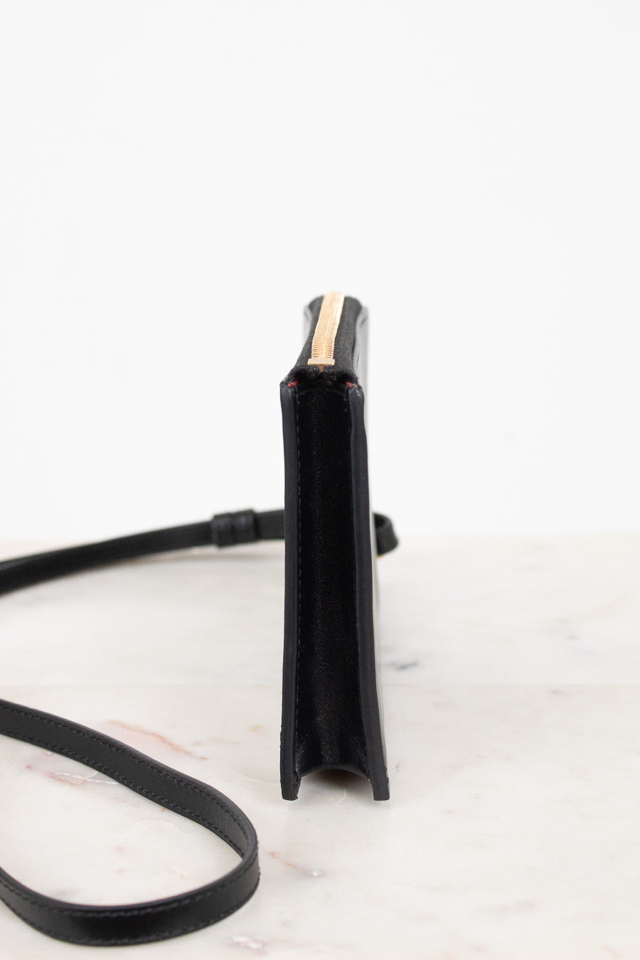 A black leather crossbody bag shown from a side profile view, featuring a gold zipper accent and a thin black strap, set against a clean white background.