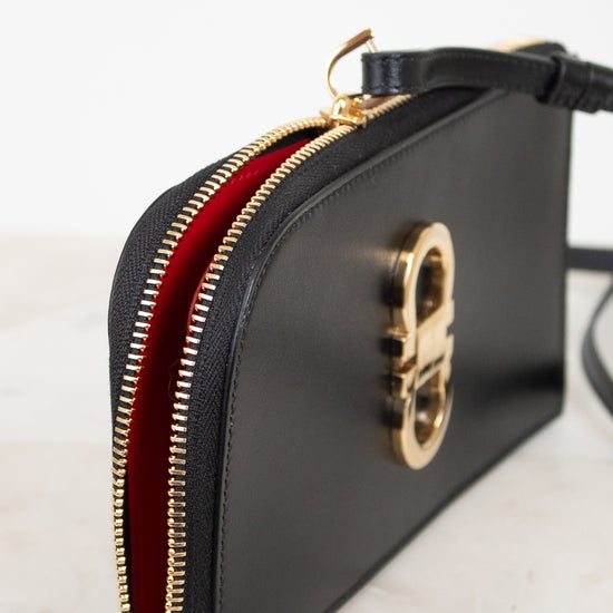 A black leather clutch bag with a gold-tone metal zipper and a vibrant red interior lining, shown on a light marble surface against a white background.