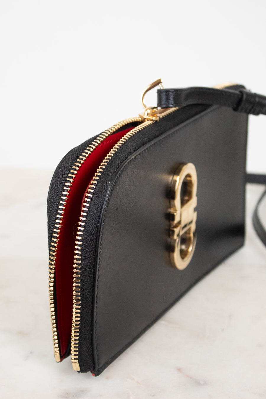 A black leather clutch bag with a gold-tone metal zipper and a vibrant red interior lining, shown on a light marble surface against a white background.