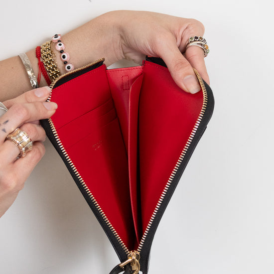A black leather zip-around wallet with a bright red interior, held open by two hands wearing gold rings and bracelets against a plain white background.