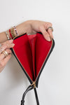 A black leather zip-around wallet with a bright red interior, held open by two hands wearing gold rings and bracelets against a plain white background.