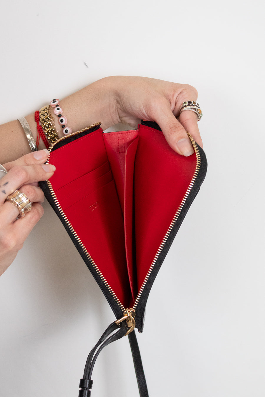 A black leather zip-around wallet with a bright red interior, held open by two hands wearing gold rings and bracelets against a plain white background.