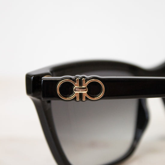 Close-up of black sunglasses with a gold-tone double-ring logo emblem on the temple arm, set against a clean white background.