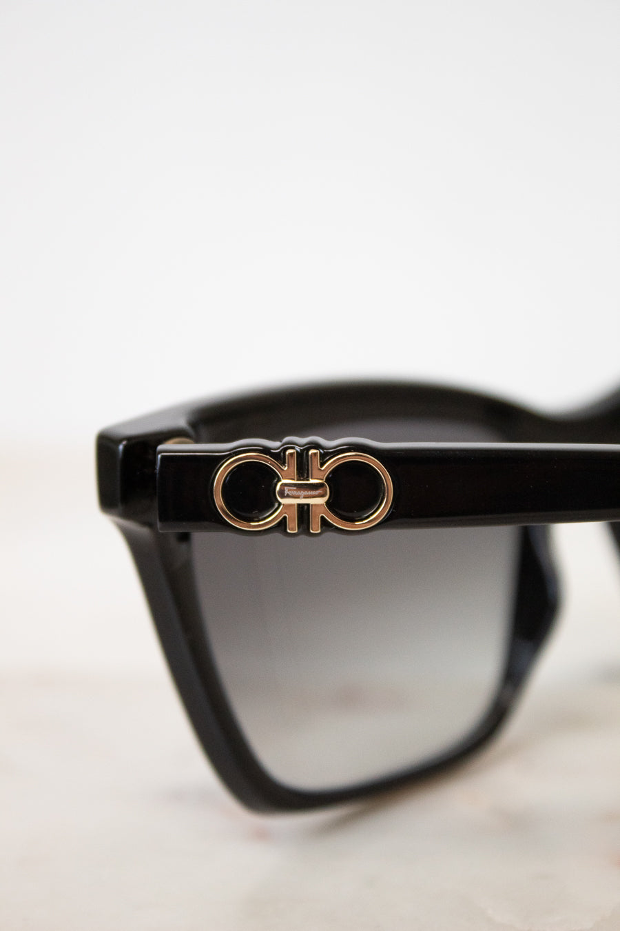 Close-up of black sunglasses with a gold-tone double-ring logo emblem on the temple arm, set against a clean white background.