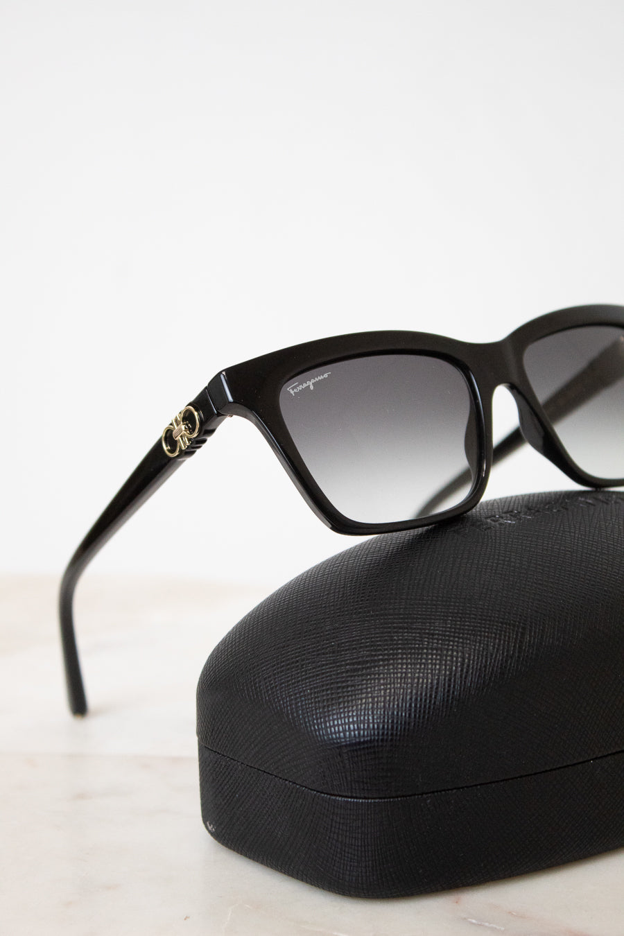 Black cat-eye sunglasses with gradient gray lenses and a gold-tone interlocking logo detail on the temple, resting on a black textured hard case against a clean white background.