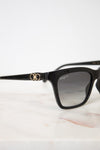 Black square sunglasses with a gold double logo on the temple arm, resting on a light marble surface against a white background.