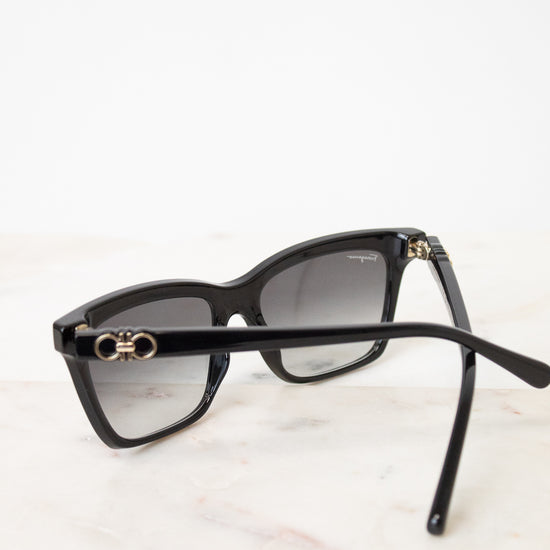 Black cat-eye sunglasses with gradient gray lenses and gold-tone double logo hardware on the temples, resting on a light marble surface.