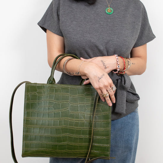 A woman wearing a gray t-shirt and blue jeans holds a green crocodile-embossed leather tote bag with a long shoulder strap against a plain white background.