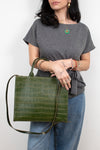 A woman wearing a gray t-shirt and blue jeans holds a green crocodile-embossed leather tote bag with a long shoulder strap against a plain white background.