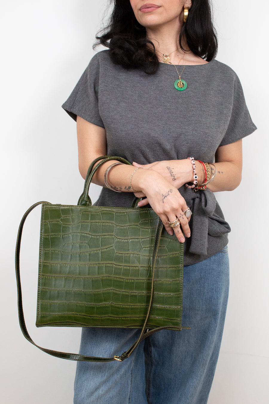 A woman wearing a gray t-shirt and blue jeans holds a green crocodile-embossed leather tote bag with a long shoulder strap against a plain white background.