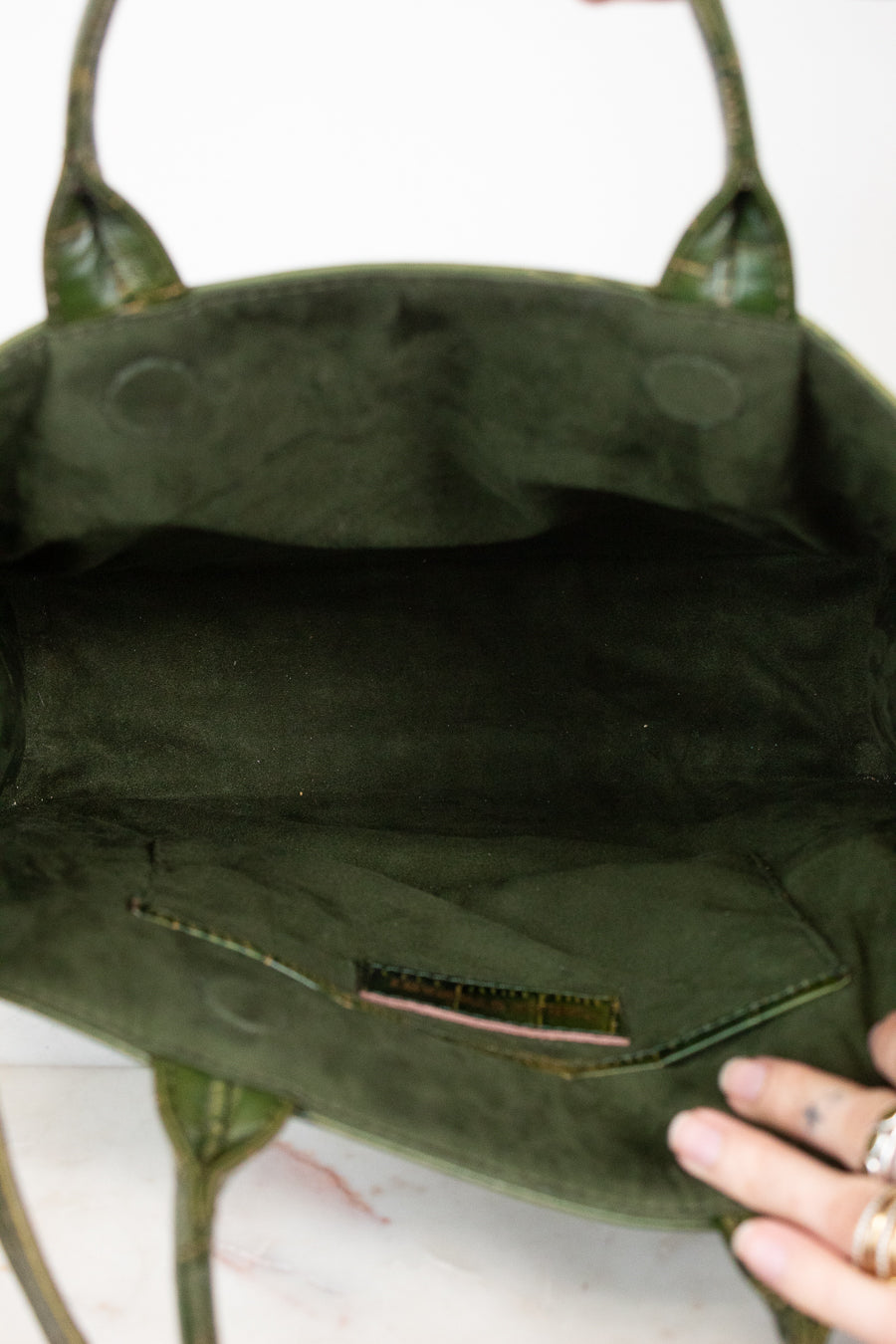 Close-up of the interior of an olive green suede handbag, showing the lining, a small interior pocket, and a hand holding the bottom edge.