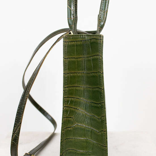 A vertical olive green crocodile-embossed leather bag with a long shoulder strap, standing on a light marble surface against a white background.
