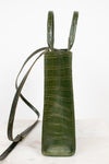 A vertical olive green crocodile-embossed leather bag with a long shoulder strap, standing on a light marble surface against a white background.