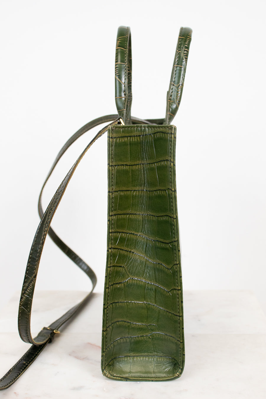 A vertical olive green crocodile-embossed leather bag with a long shoulder strap, standing on a light marble surface against a white background.