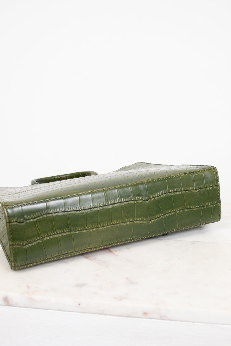 An olive green crocodile-embossed leather clutch bag resting on a white marble surface against a white background.