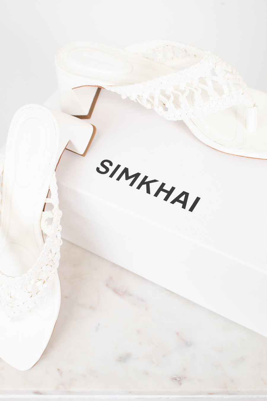 White block heeled sandals with white box branded 'SIMKHAI' on a marble surface