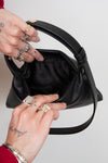 Person holding a black handbag with tattoos and jewelry on fingers against a plain background
