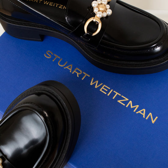 Black loafers with decorative charm on a blue Stuart Weitzman box.