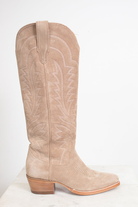 A single knee-high western boot in light tan suede with white embroidered stitching, shown in profile against a plain white background.