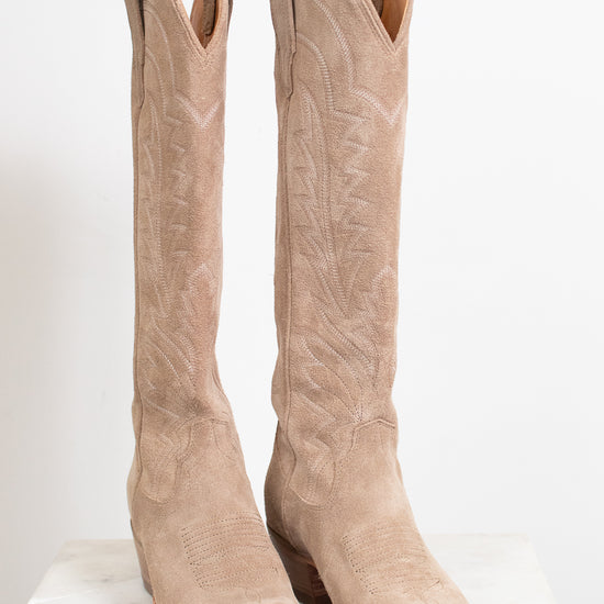 A pair of tall western boots in light tan suede with decorative stitching, displayed on a white marble surface against a white background.