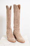 A pair of tall western boots in light tan suede with decorative stitching, displayed on a white marble surface against a white background.