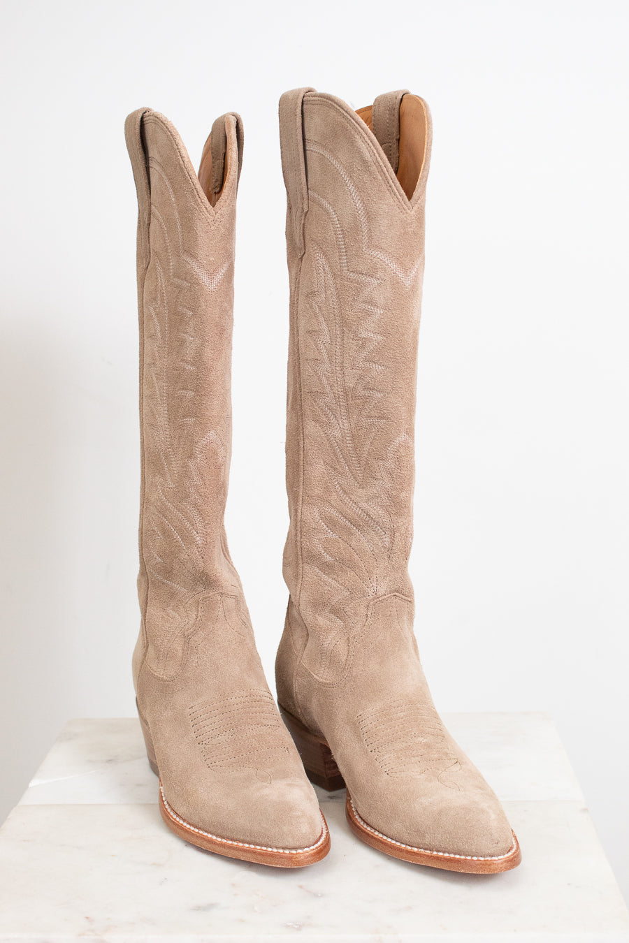 A pair of tall western boots in light tan suede with decorative stitching, displayed on a white marble surface against a white background.