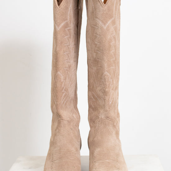 A pair of tall western boots in light tan suede with white embroidered stitching, displayed on a white marble surface against a white background.