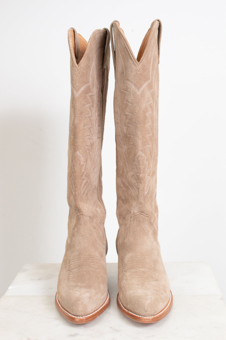 A pair of tall western boots in light tan suede with white embroidered stitching, displayed on a white marble surface against a white background.