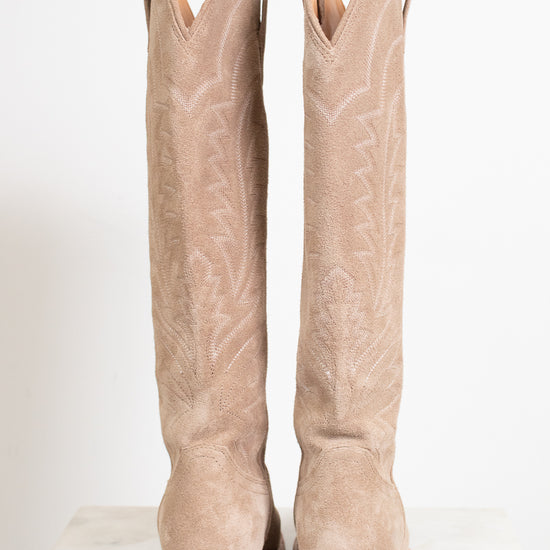 A pair of tall western-style boots in light tan suede, shown from the back on a white marble surface against a white background.